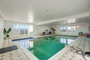 An indoor swimming pool with clear water, surrounded by white walls, large windows, and a ceiling with recessed lighting. - Bay Area Family Home w/ Indoor Pool + Game Room (South San Francisco)