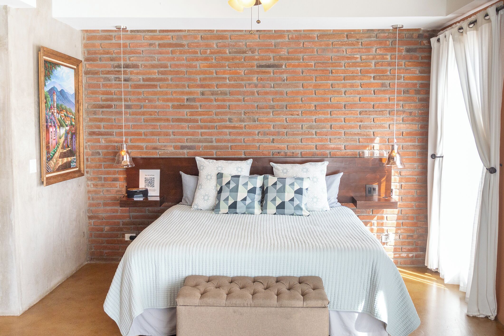 A bedroom with a brick wall, a bed with a white bedspread, two decorative pillows, a wooden headboard, a bedside table with a lamp, and a painting on the wall.