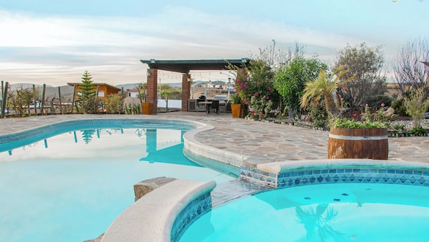Una piscina con solárium de piedra, un patio cubierto y jardines aledaños.