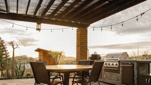 Un patio al aire libre con mesa de piedra, sillas de mimbre, una parrilla y luces colgantes.
