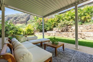 A covered outdoor seating area with a sofa and table, surrounded by lush greenery and a view of a mountainous landscape. - @ Marbella Lane Hale Lai Serene Luxury 3BR Home (Waianae)