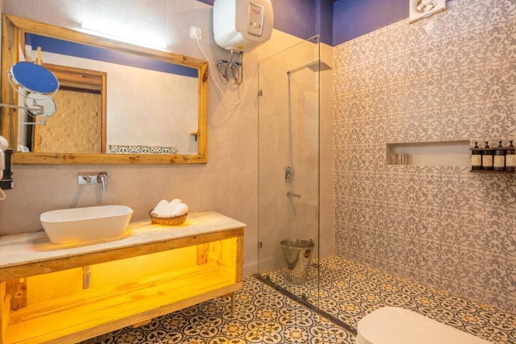 A bathroom with a wooden vanity, a white sink, a mirror, a shower, patterned tiles, and a shelf with bottles.