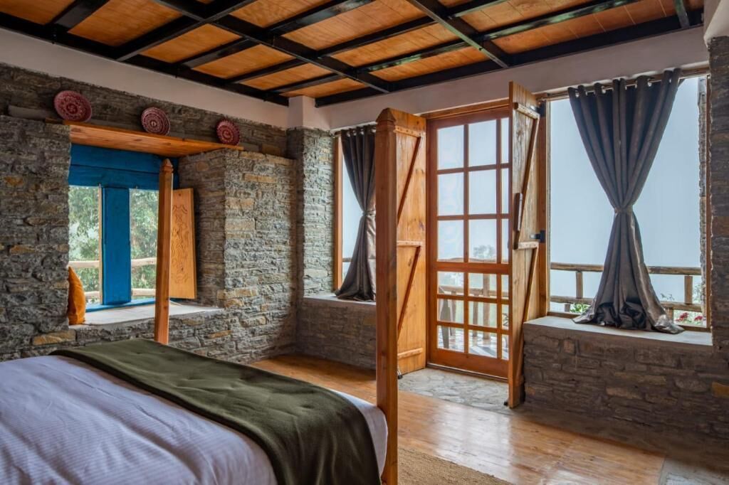 A rustic room with a bed, stone walls, wooden beams, and large windows with curtains.