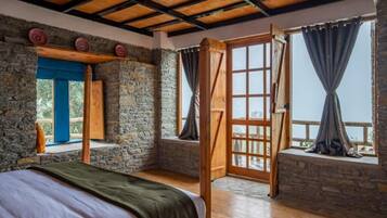 A rustic room with a bed, stone walls, wooden beams, and large windows with curtains.