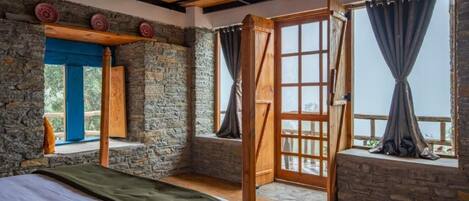 A rustic room with a bed, stone walls, wooden beams, and large windows with curtains.
