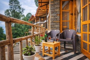 Ein Holzbalkon mit Schachbrettfußboden, zwei Stühle, einem Tisch mit Flasche und Gläsern sowie Topfpflanzen.
