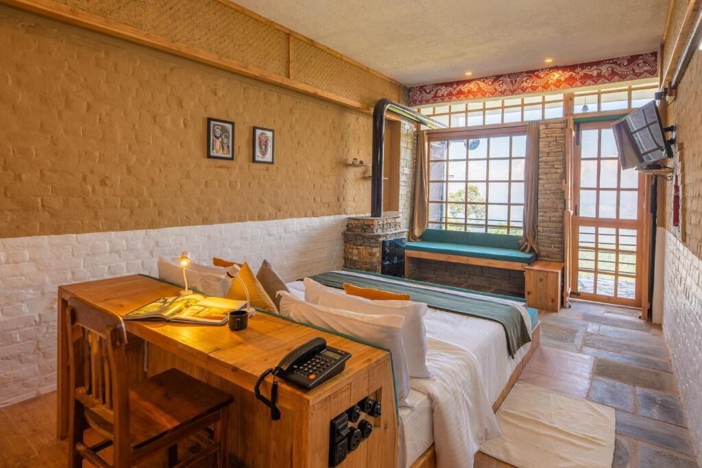 Ein Zimmer mit einem Holztisch, einem Bett mit grünem Kopfteil, einer Steinwand und einem großen Fenster.
