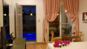 Una habitación con una cama, un escritorio con computadora, una silla, una pequeña mesa con flores y una puerta corrediza de vidrio que da a una zona de piscina.