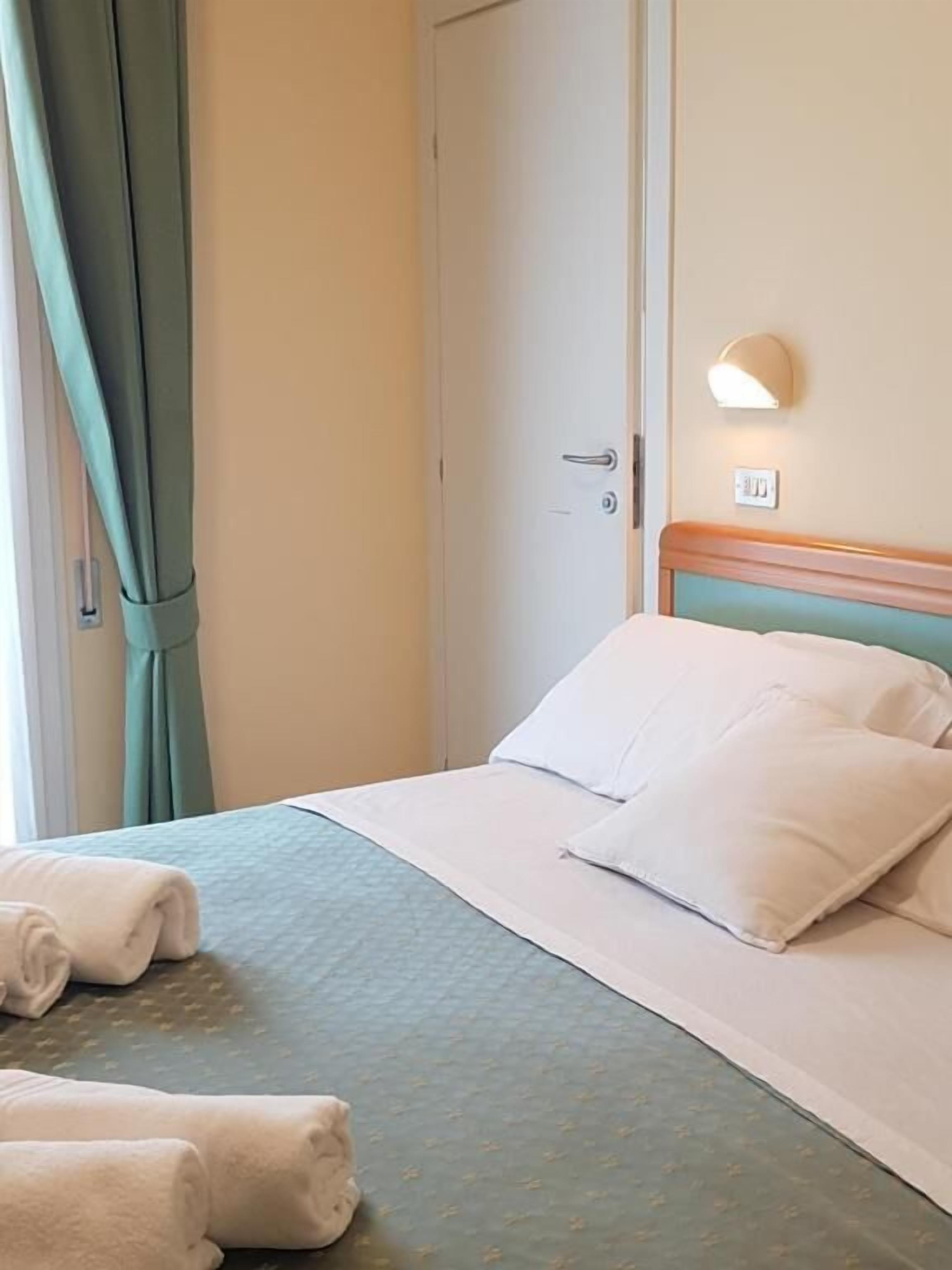 Una camera d'albergo con un letto, cuscini, una testiera, una luce a parete e una porta.