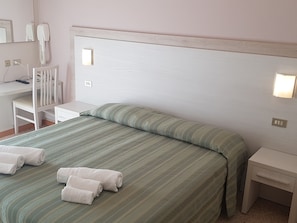 A neatly made bed with green bedding and white towels, a white desk with a chair, and bedside lamps.
