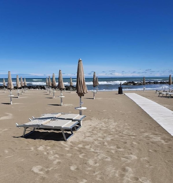 A beach with rows of sun loungers and umbrellas, a concrete walkway, and the ocean in the background.