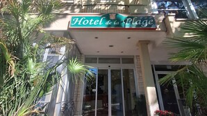 Entrada de un hotel con un letrero que dice 'Hotel de la Plage', una puerta de cristal y un pequeño espacio al aire libre con mesas y sillas.