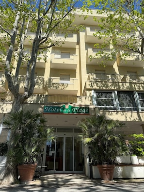 A hotel facade with a sign reading 'Hotel de la Plage,' large potted plants, and a multi-story building with balconies.