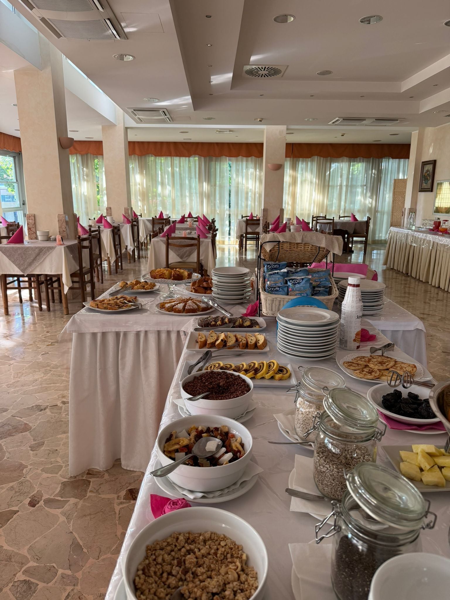 Ein reichhaltiges Buffet mit verschiedenen Speisen, Tischen und Stühlen in einem hellen Raum mit großen Fenstern.