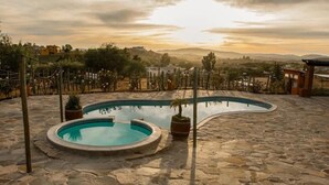 Uma piscina de pedra com borda curva, rodeada por um terraço de pedra, e vista para colinas e edifícios ao longe.