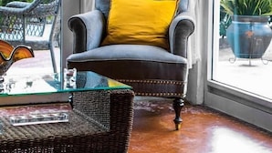 Um quarto com uma janela grande, uma poltrona estofada cinza com almofada amarela, uma cadeira de vime e uma mesa de centro de vidro.