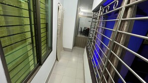 Un couloir étroit avec une rambarde en métal, une fenêtre avec des stores horizontaux et un sol carrelé.