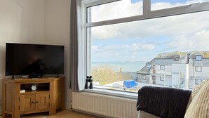 A room with a large window, a flat-screen TV on a wooden stand, a radiator, and a view of buildings and water. - Clearwater Seaview a perfect retreat (St Ives)