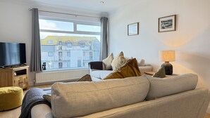 A living room with a sofa, a TV on a wooden stand, a lamp, and a window with a view of buildings. - Clearwater Seaview a perfect retreat (St Ives)
