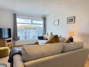 A living room with a sofa, a TV on a wooden stand, a lamp, and a window with a view of buildings. - Clearwater Seaview a perfect retreat (St Ives)