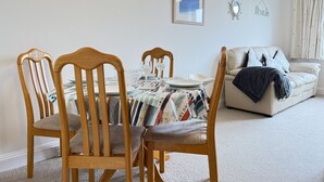 Uma área de refeições com uma mesa posta para quatro pessoas, cadeiras de madeira, toalha de mesa estampada e um sofá ao fundo.