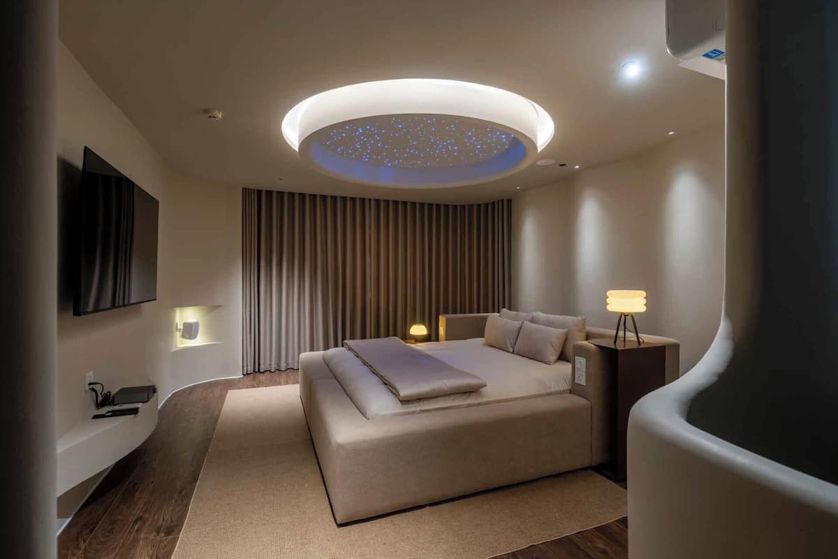 Ein modernes Schlafzimmer mit einem großen Bett, einer Deckenleuchte, einem Nachttisch und einem an der Wand montierten Fernseher.