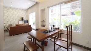 A modern living room with a wooden dining table, a sofa, and a large window with a view of greenery. - Palette Amura Hotel - Near Gigamall (Ho Chi Minh City)