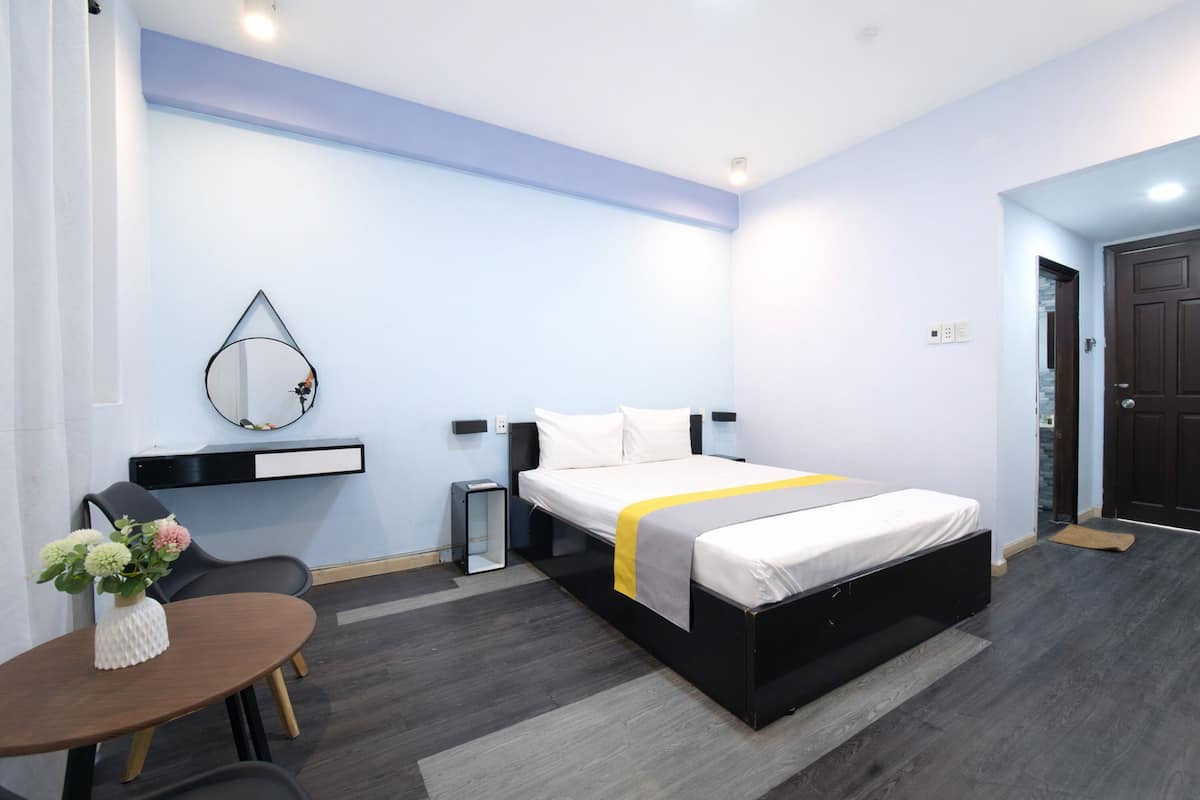 A modern hotel room with a bed, bedside tables, a mirror, a small table with a vase, and a door.