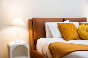 Un lit bien fait, avec des oreillers blancs et moutarde, une table de chevet blanche avec une lampe, et une tête de lit marron.