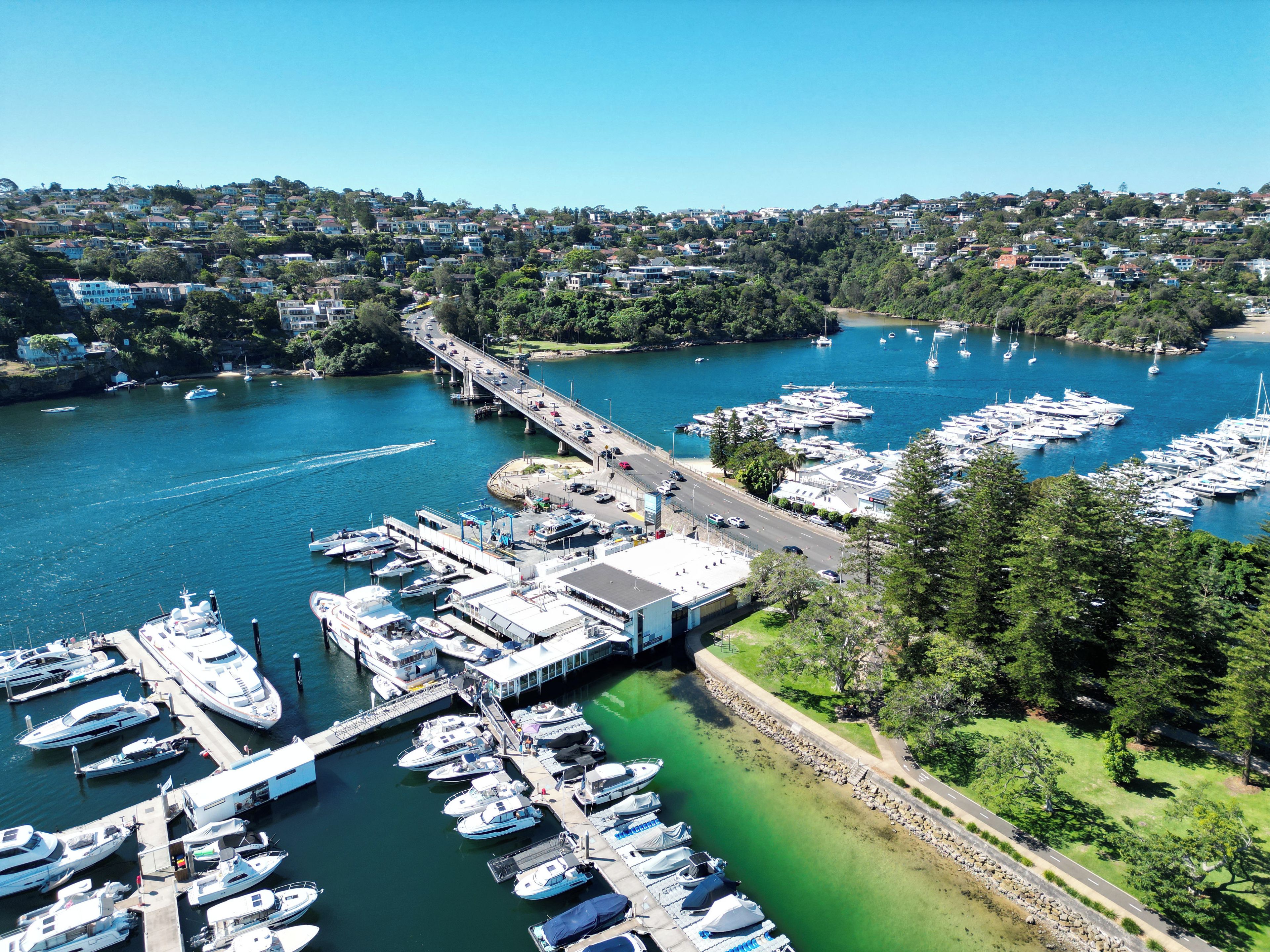 A marina with multiple boats docked, a bridge connecting to the land, and a residential area in the background.
