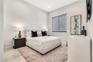 Ein modernes Schlafzimmer mit einem großen Bett, Nachttischen, einem Fenster mit Jalousien und einem abstrakten Kunstwerk an der Wand.