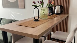 A dining area with a wooden table, four grey chairs, a black vase with flowers, a kettle, and a wall-mounted abstract painting.