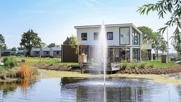 Ett modernt hus med en fontän i en damm, omgivet av en välskött gräsmatta och andra hus.