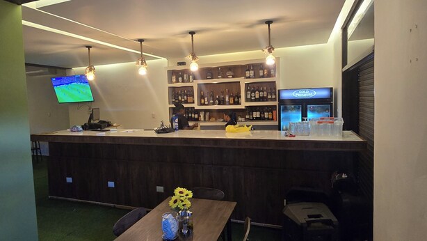 A bar area with a counter, shelves of liquor bottles, a television, and a table with a vase of flowers.