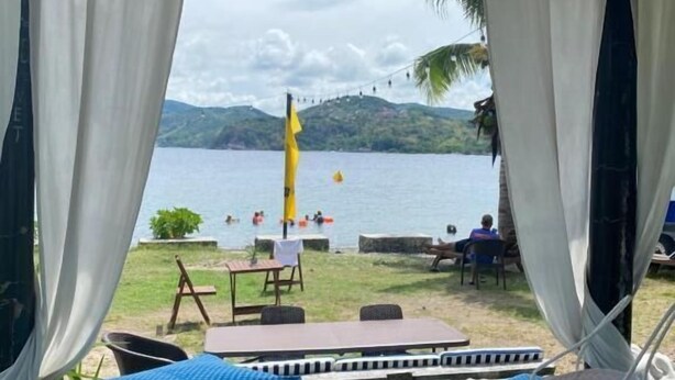 Une vue sur la plage avec une structure dotée d’un toit de chaume, une table, des chaises et un tapis agrémenté de coussins colorés.