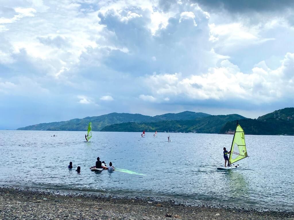 A beach with people windsurfing, calm water, and distant mountains.