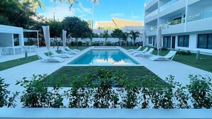 Ein Poolbereich mit weißen Liegestühlen und einem rechteckigen Pool, umgeben von Palmen und einem modernen Gebäude mit Balkonen.
