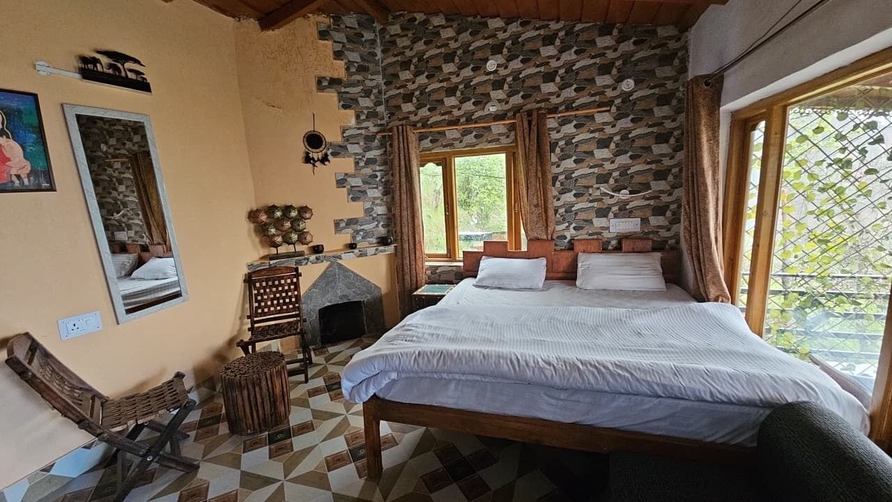 A bedroom with a stone wall, a bed with white linens, a wooden bench, a small fireplace, and a window with curtains.
