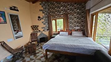 A bedroom with a stone wall, a bed with white linens, a wooden bench, a small fireplace, and a window with curtains.