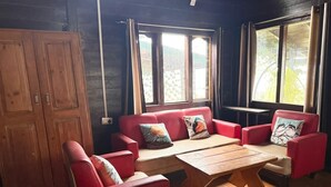 Een kamer met een houten vloer, een rode zithoek, een houten tafel en grote ramen met gordijnen.