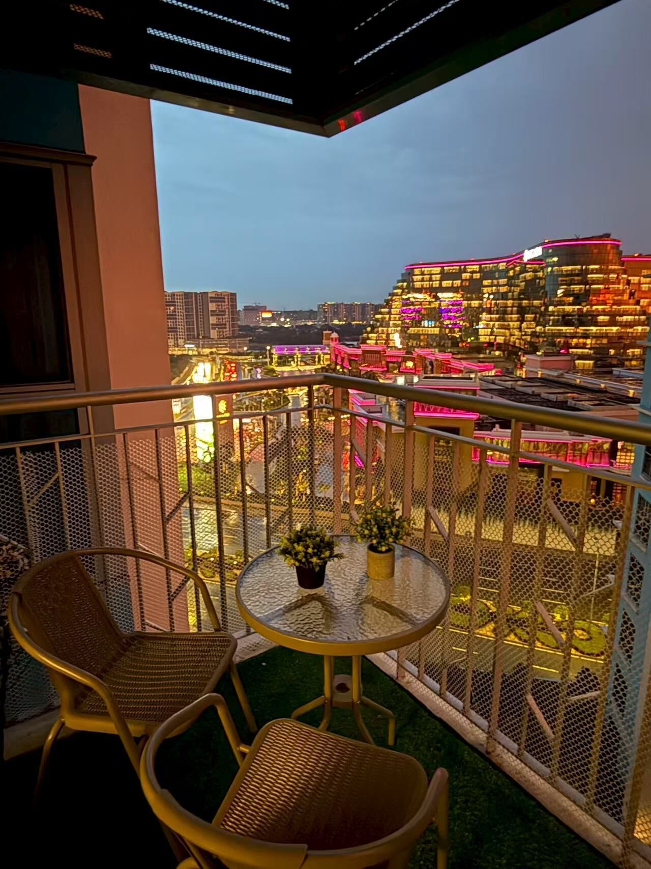 Un balcón con mesa y sillas, vista a un paisaje urbano con edificios iluminados, y barandilla de malla.