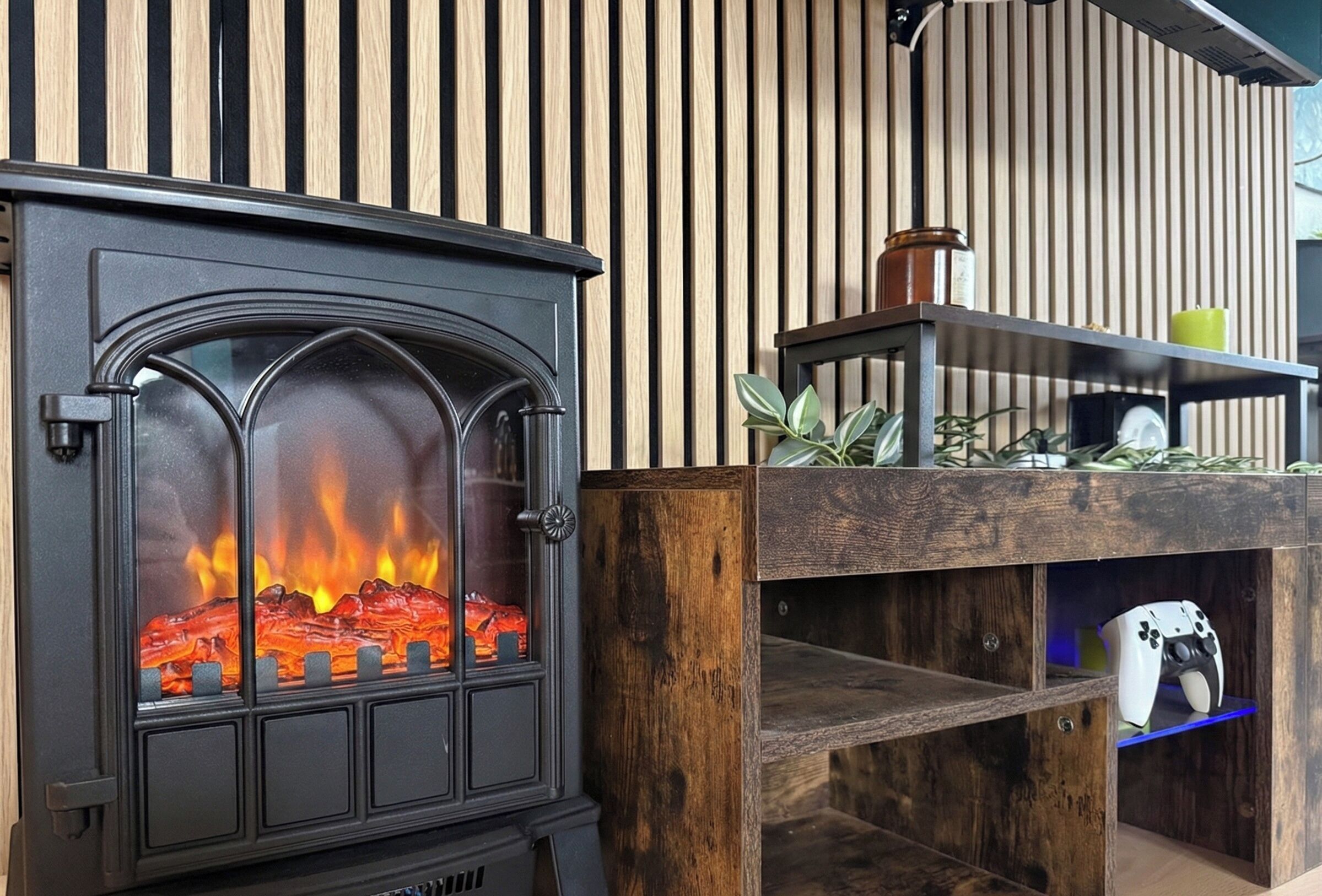 A wood-burning stove with a glass door, a shelf with a candle, and a gaming console in a wooden cabinet.