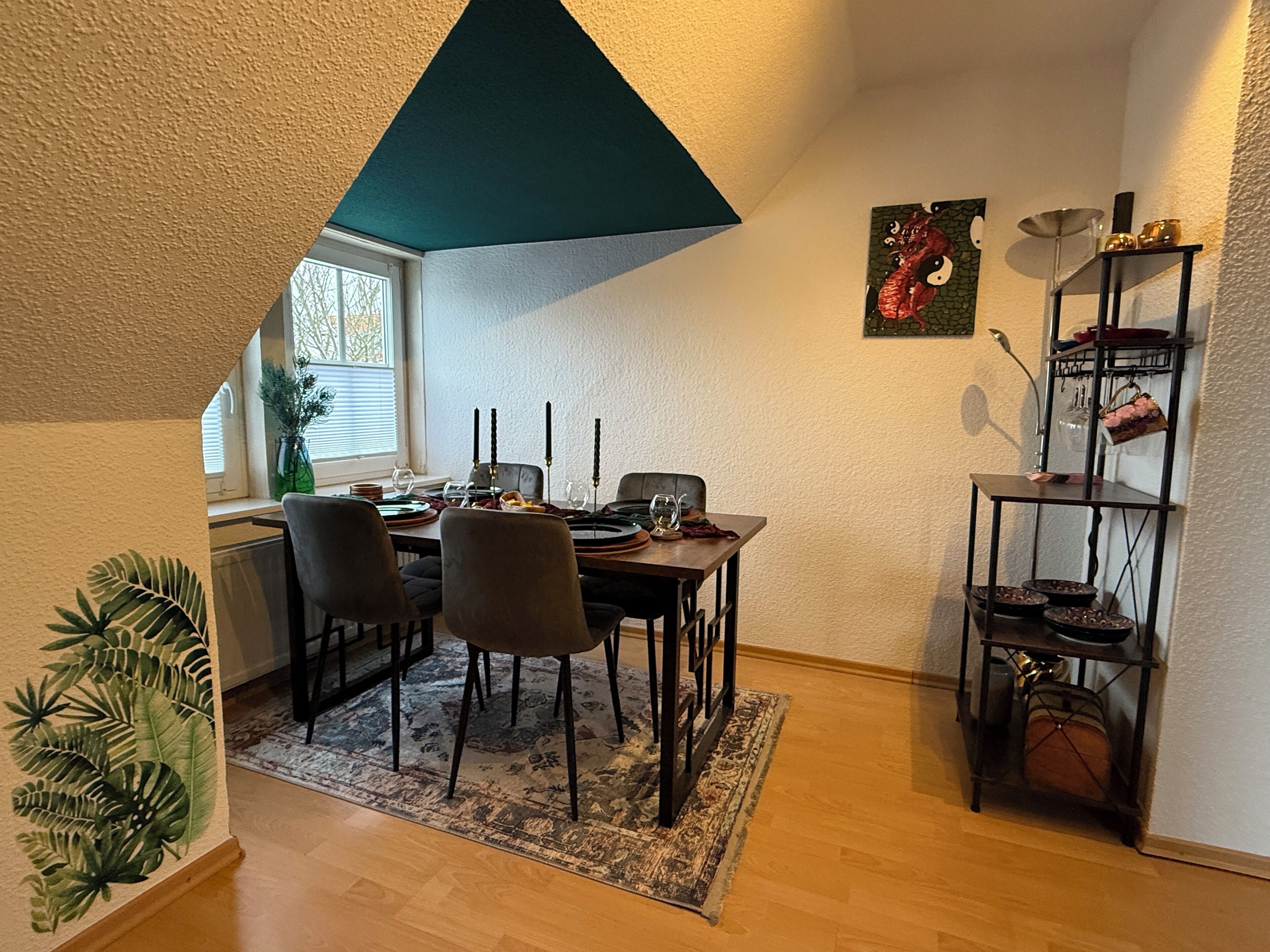 A dining area with a table set for four, chairs, a rug, a window with blinds, and a decorative shelf with items.