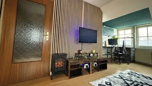 Una sala de estar con chimenea, televisor montado en la pared, escritorio con computadora y ventana con persianas.