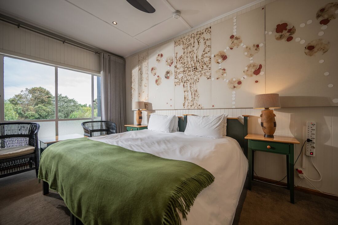 A hotel room with a large bed, a green throw blanket, a chair, a nightstand with a lamp, and a wall with a decorative pattern.