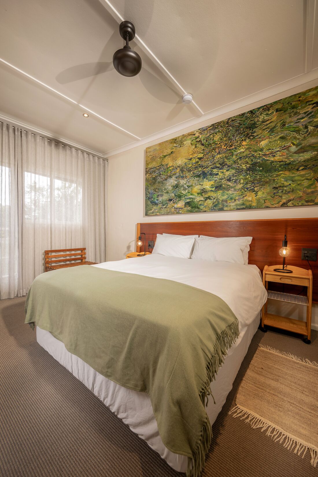 Habitación de hotel con cama, ventilador de techo, mesita de noche con lámpara, un cuadro en la pared y ventana con cortinas.