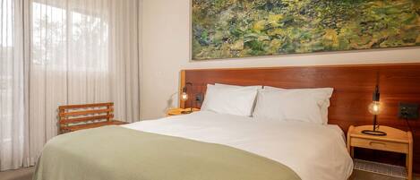 Habitación de hotel con cama, ventilador de techo, mesita de noche con lámpara, un cuadro en la pared y ventana con cortinas.