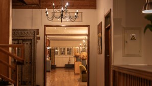 Un couloir orné d’un lustre, avec une porte en bois, donnant sur une pièce où l’on aperçoit une lampe et des tableaux encadrés.
