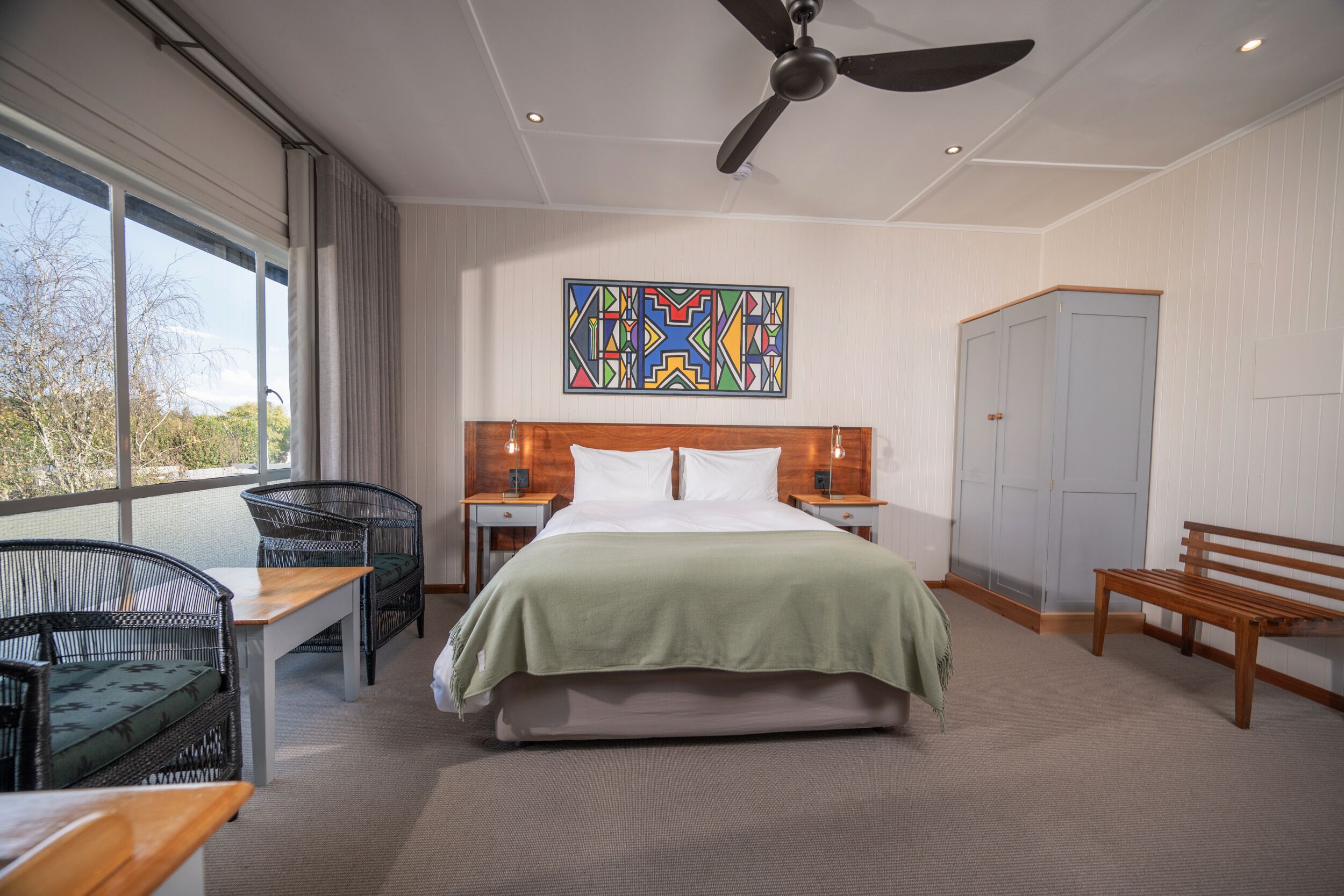 A hotel room with a bed, a desk, two chairs, a bench, a wardrobe, a ceiling fan, and a window with a view.