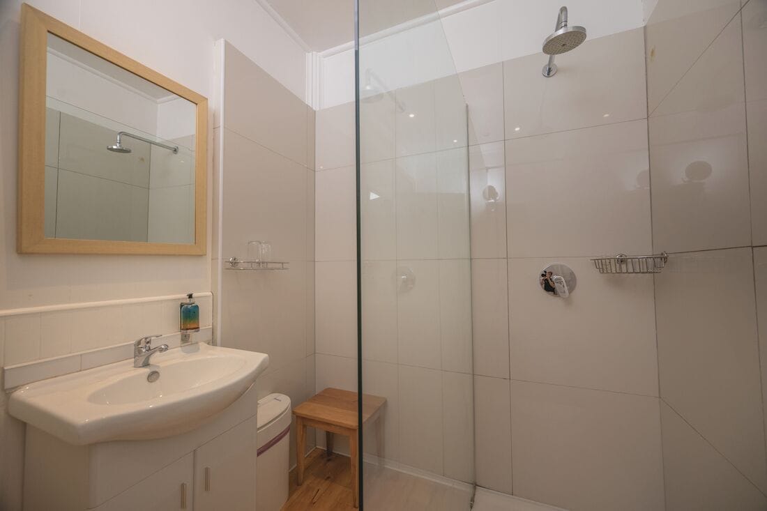 Ein Badezimmer mit Duschkabine, Waschbecken, Spiegel und einem kleinen Holzschemel.